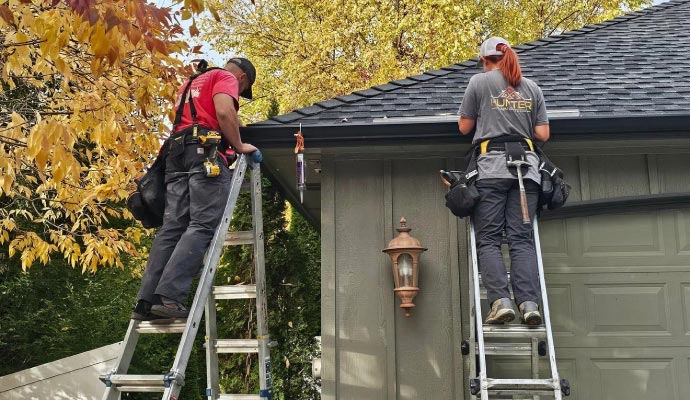 Two technicians repairing residential gutters while standing on ladders, fixing leaks and alignment issues to restore proper drainage.