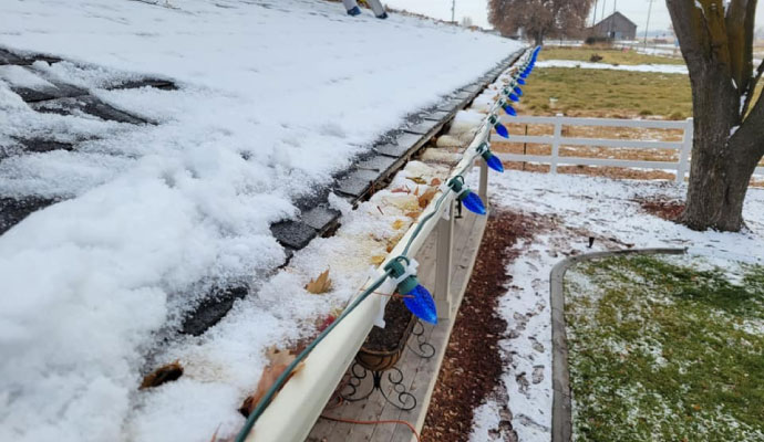 Snow-covered roof gutter with string lights along the edge of a house